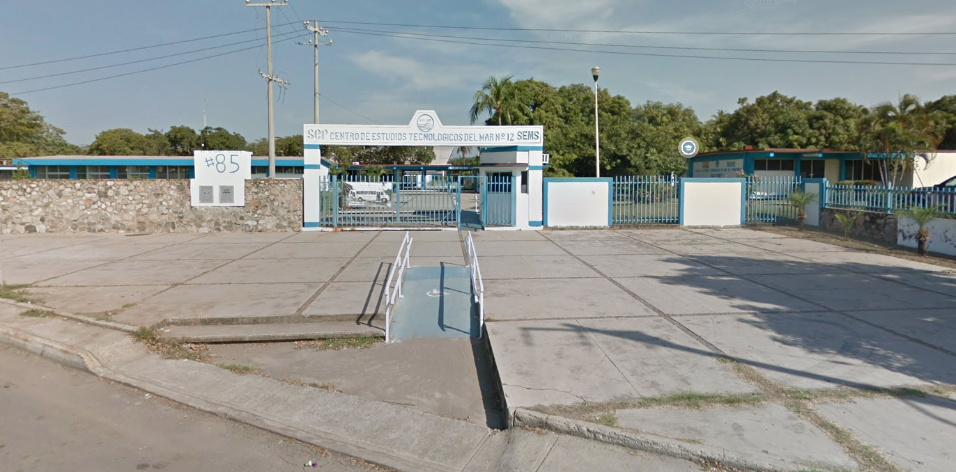 Fotografía en la que se muestra una rampa que da acceso a una explanada. Al fondo de la explanada se encuentra la entrada del plantel conformada por una reja de color azul y arriba de ella, en letras azules dice CENTRO DE ESTUDIOS TECNOLÓGICOS DEL MAR, 12. 